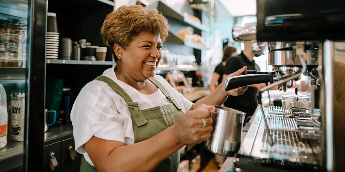 2025-06-Junio01_EconomiaPlateada_agewithoutlimits Una mujer mayor sonríe mientras prepara una bebida cliente en el negocio que atiende