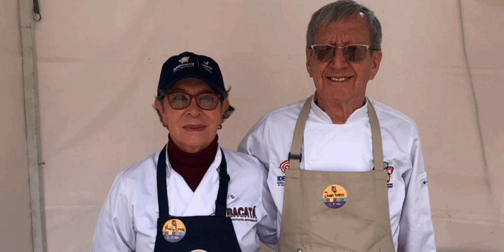 Emma, mujer de 72 años de edad, y Juan, hombre de 74 años de edad, en una feria de emprendimiento. Ella viste un delantal azul y él, uno camel.