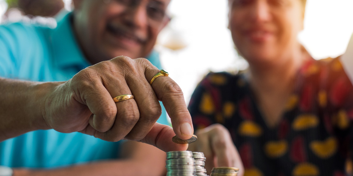 Sin título-2 Un hombre mayor acomoda cuidadosamente unas monedas sobre una mesa, mientras a su lado una mujer mayor lo observa con atención. Ambos están sentados.