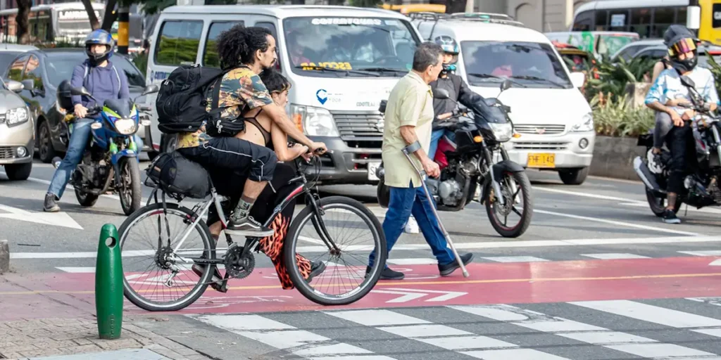 Se observan diferentes actores viales en el cruce de una avenida de alta circulación: una persona atravesando un paso peatonal en muleta, un joven en bicicleta y colectivos, moticicletas y carros particulares, en la ciudad de Medellín