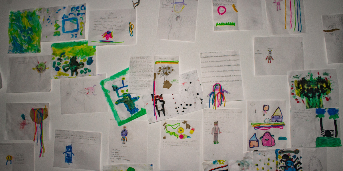 Fotografía de una pared blanca completamente cubierta por hojas de papel de distintos colores pegadas unas al lado de otras. En cada hoja hay poemas escritos a mano por niños. Los textos son breves, algunos están acompañados de pequeños dibujos. La pared se ve desordenada pero llena de vida, como un gran collage de palabras e infancia.