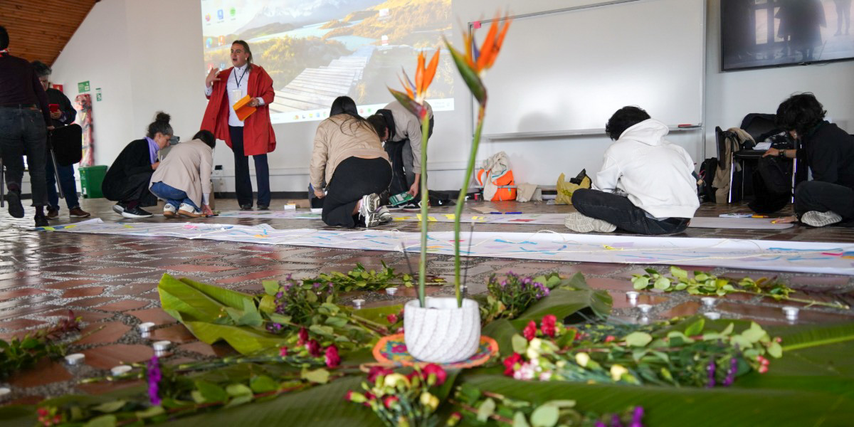 Grupo de personas adultas sentadas en c&iacute;rculo en un taller. Sobre la mesa hay flores de colores y materiales de trabajo. Algunas personas r&iacute;en mientras otras recortan y organizan las flores.