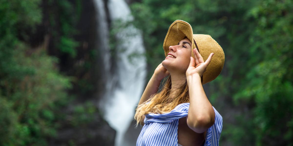 Imagen generada con Inteligencia Artificial de una mujer de perfil joven que mira un gran paisaje de cascada. Lleva ropa c&oacute;moda de turista, sombrero.