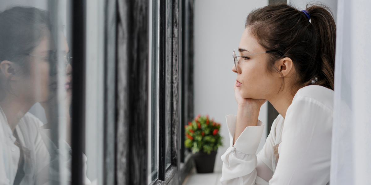 Mujer mayor sentada junto a una ventana, mirando hacia afuera con expresi&oacute;n pensativa y melanc&oacute;lica, reflejada en el vidrio.