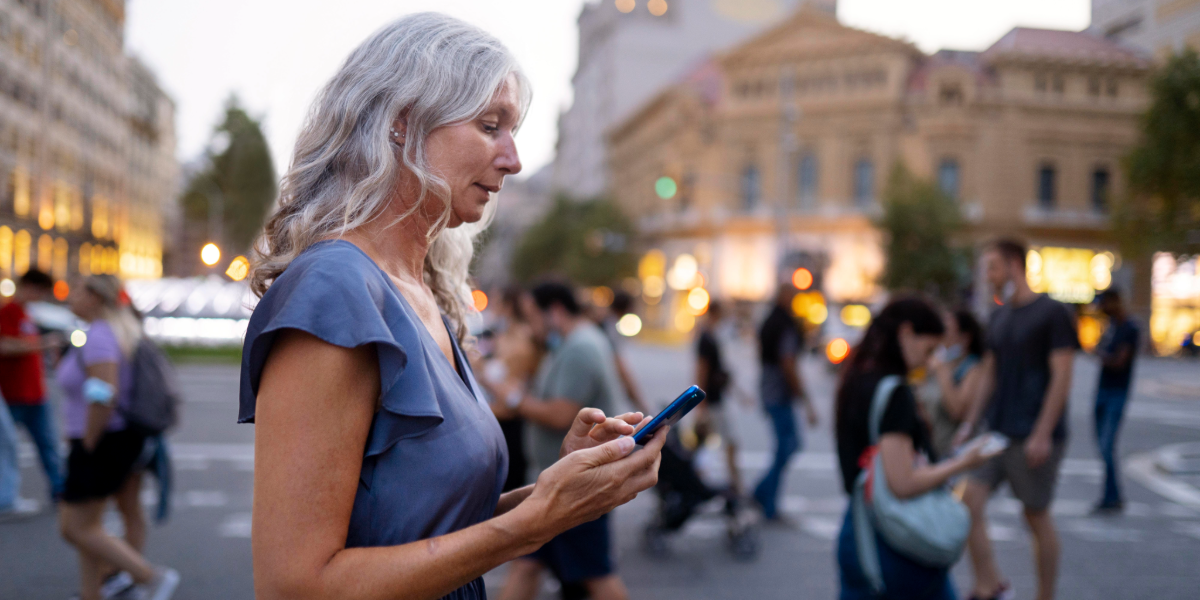 Mujer de aproximadamente 60 a&ntilde;os, cabello largo y ropa casual, camina por una acera de una ciudad mirando fijamente la pantalla de su tel&eacute;fono celular que sostiene con una mano. Detr&aacute;s y a los costados, grupos de personas caminando. Al fondo se ven edificios urbanos y una calle con tr&aacute;nsito. La imagen transmite la paradoja de estar rodeado de gente pero aislado en la propia burbuja digital.